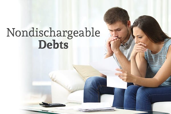 A husband and wife sit on a couch looking with concern at a paper she's holding. A stack of other papers and a calculator sit on the coffee table before them. There's also the words "Nondischargeable Debts"